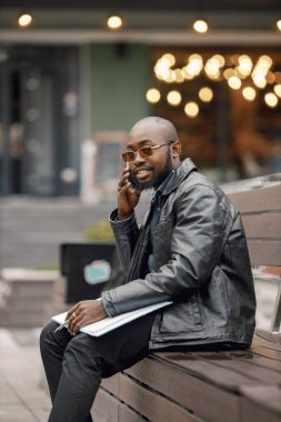 Şehirdeki bir bankta oturan siyah bir adam. Güneş gözlüklü ve deri ceketli bir adam. İş adamının evrakları var ve telefon kullanıyor..