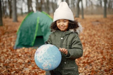 Ormanda kamp yapan küçük siyah bir kız. Kız elinde bir küre tutuyor. Haki ceket ve bej şapka giyen siyah bir kız..