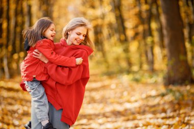 Young woman with little girl walking in autumn forest. Blonde woman play with her daughter and holding her on a hands. Mother and daughter wearing jeans and red jackets.