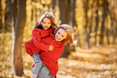 Young woman with little girl walking in autumn forest. Blonde woman play with her daughter and holding her piggyback. Mother and daughter wearing jeans and red jackets.