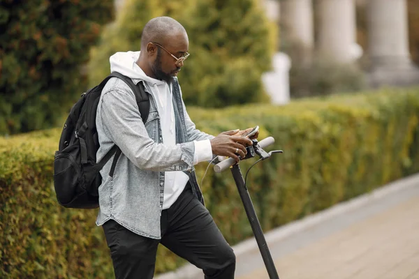 Elektrikli scooterlı siyah bir adam sokakta dikiliyor. Beyaz kapüşonlu ve siyah kotlu bir adam. Telefon kullanan siyah çantalı bir adam..