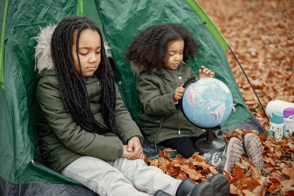 Beautiful little black girls in tent camping in the forest. Two little sisters sitting in a tent in autumn forest and looking on a globe. Black girls wearing khaki coats.