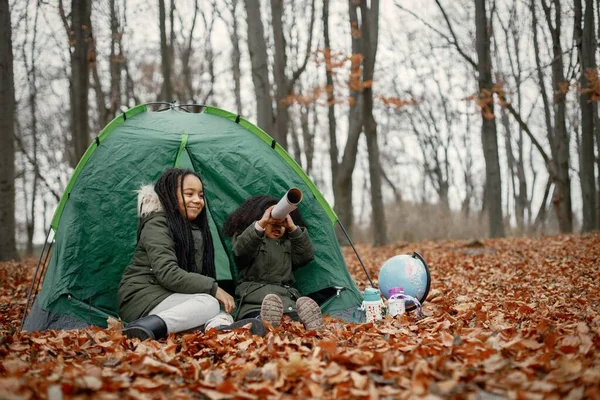 Beautiful little black girls in tent camping in the forest. Two little sisters sitting in a tent in autumn forest and looking on a globe. Black girls wearing khaki coats.