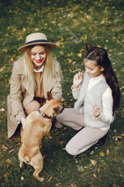 Kadın, küçük kız ve köpek sonbahar parkında oynuyorlar. Kahverengi buldog ve bayan sahipleri bir gün parkta oyun oynayıp eğleniyorlar. Bej ceketli ve şapkalı bir kadın..