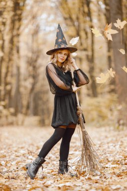 Blonde young girl witch in forest on Halloween. Girl wearing black dress and cone hat. Witch holding a broom.