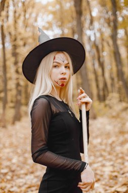 Blonde young girl witch in forest on Halloween. Girl wearing black dress and cone hat. Witch holding a broom.