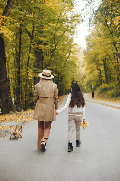Woman, little girl and dog walking in the autumn park. Brown french bulldog with female owners spend a day at the park playing and having fun. Woman wearing beige coat and a hat.