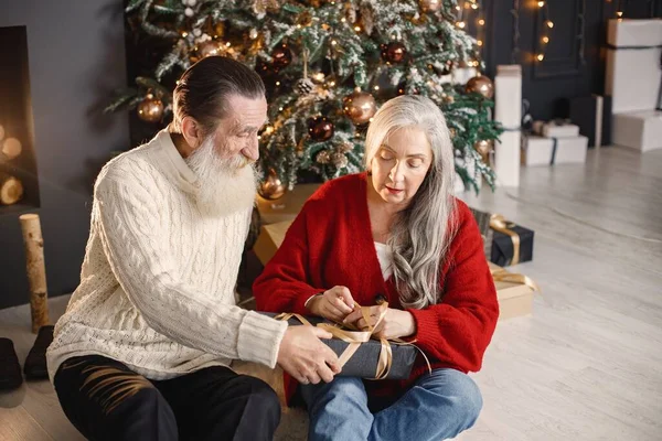 Senior man celebrating christmas with his wife near beautiful christmas tree. Old bearded man sitting on a floor near his wife with grey hair and holding a gifts. Man wearing white sweater and woman