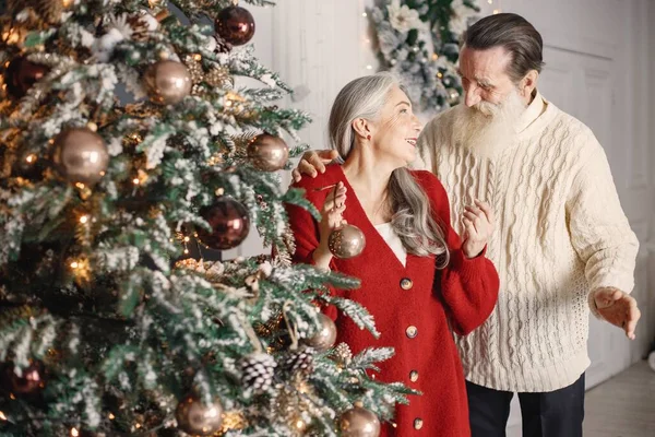 Senior man celebrating christmas with his wife near beautiful christmas tree. Old bearded man standing near his wife with grey hair. Man wearing white sweater and woman red one.