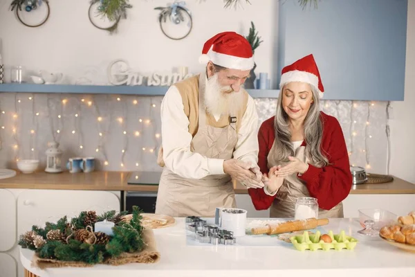 Senior man and his wife cooking together. Old bearded man standing together near his wife with grey hair. Man wearing white sweater and woman red one.