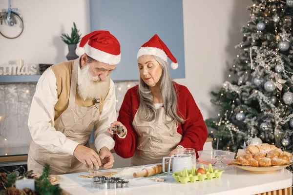 Senior man and his wife cooking together. Old bearded man standing together near his wife with grey hair. Man wearing white sweater and woman red one.