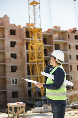 İnşaat alanı yöneticisi çelik yelek ve kask giyip inşaat alanında düşünüyor. İnşaat alanında evrakları olan bir mimar. Telefonda konuşan adam..