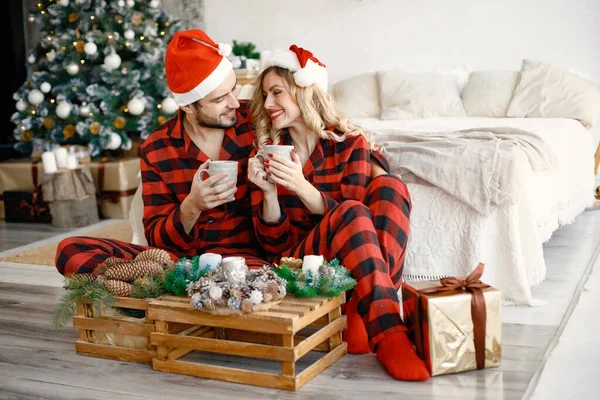 Romantik çift Noel 'i Noel ağacının yanında yerde kutluyor. Sarışın kadın ve ekose pijama giyen esmer adam. Kupalardan çay içen romantik bir çift..