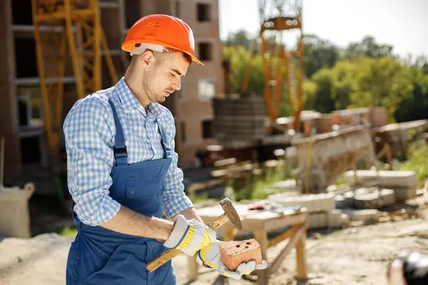 Worker standing wearing orange helmet. Man holding a hammer and a brick in his hands. Construction site on a background.