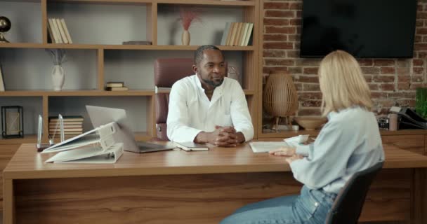 白い医療服の黒人医師は 私立病院で女性患者と相談しています 男性医師 診療所での会議で女性のクライアントに相談 — ストック動画