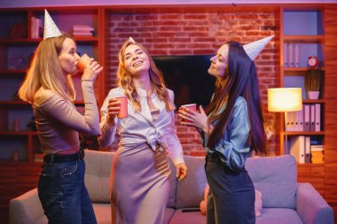 Genç arkadaşlar akşam partisinde evde dikiliyorlar. Herkes parti konisi takıyor. Elinde içeceklerle kağıt bardak tutan kızlar..