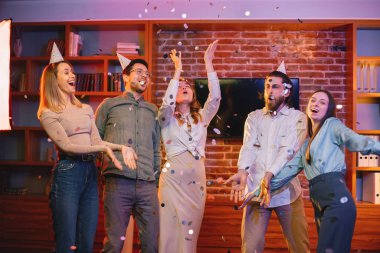 Genç arkadaşlar akşam partisinde evde dikiliyorlar. Erkekler ve kızlar konfeti altında eğleniyorlar. Herkes parti konisi takıyor..