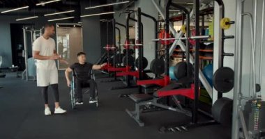 Tekerlekli sandalyedeki engelli adam koçla çalışıyor. Rehabilitasyon uzmanı, tekerlekli sandalyedeki bir adama egzersiz yapması için yardım ediyor..