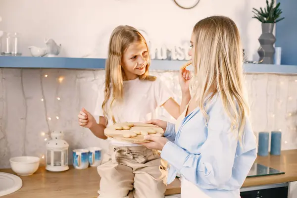 Family preparation of ginger biscuits with daughter. Mother and little girl trying a cookies. Blonde woman wearing blue shirt and girl white t-shirt.
