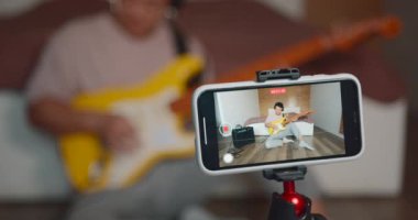 Gitar çalan mutlu genç adam, akıllı telefon web kamerasında kendi ses performansını kaydediyor. Profesyonel blog öğretmeni çevrimiçi canlı video yayınlıyor.