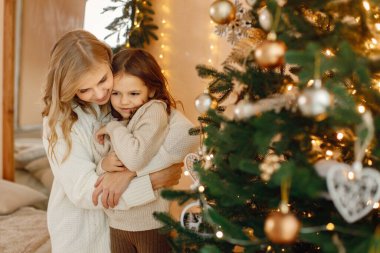 Noel 'i annesiyle birlikte kutlayan küçük bir kızın portresi. Kız ve annesi Noel ağacının yanında dikilirken eğleniyorlar. Bej örgülü giysiler giyen bir aile..