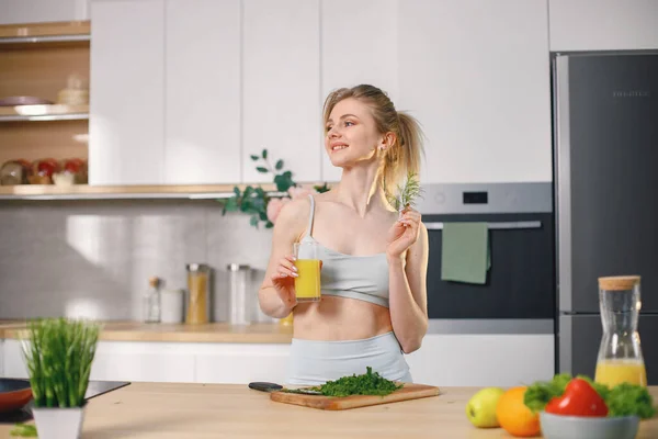 Fit woman with lots of healthy fresh food at home. Woman in grey top drinking a glass with juice. Concept of losing weight, sports and healthy eating.
