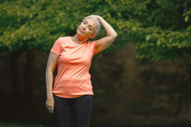 Park 'ta dikilen gül tişörtlü son sınıf öğrencisi kadın alıştırmalar yapıyor. Olumlu olgun kadınlarda fitness ya da yoga dersi vardır. Emeklilikte sağlık ve sağlıklı yaşam tarzı.