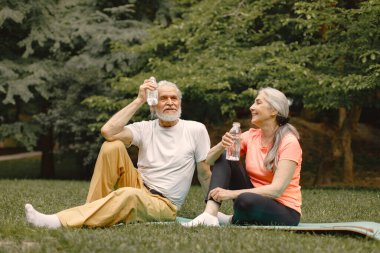 Yoga minderli yaşlı adam ve kadın ellerinde su şişeleriyle parkta oturuyorlar. Olgun bir adam ve yaşlı karısı spor yaptıktan sonra ya da yoga dersinden sonra. Emeklilikte sağlık ve sağlıklı yaşam tarzı.