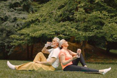 Yoga minderli yaşlı adam ve kadın ellerinde su şişeleriyle parkta oturuyorlar. Olgun bir adam ve yaşlı karısı spor yaptıktan sonra ya da yoga dersinden sonra. Emeklilikte sağlık ve sağlıklı yaşam tarzı.