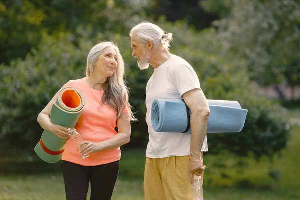 Senior man and woman with yoga mats standing at park. Positive mature man and his elderly wife after fitness or yoga class in nature. Wellness and healthy lifestyle on retirement.
