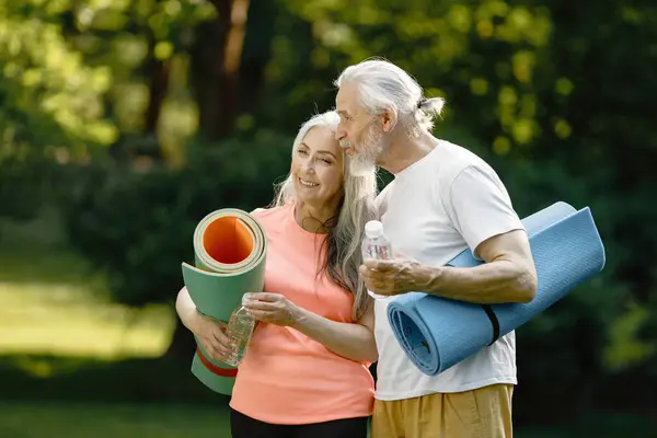 Senior man and woman with yoga mats standing at park. Positive mature man and his elderly wife after fitness or yoga class in nature. Wellness and healthy lifestyle on retirement.