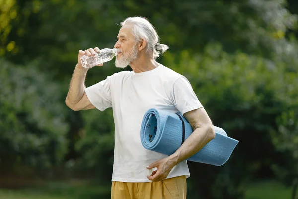 Senior man with yoga mat standing at park with bottle of water. Positive mature man after fitness or yoga class in nature. Wellness and healthy lifestyle on retirement.