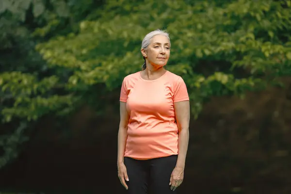 Senior woman in rose t-shirt standing at park and doing excercises. Positive mature woman has fitness or yoga class in nature. Wellness and healthy lifestyle on retirement.