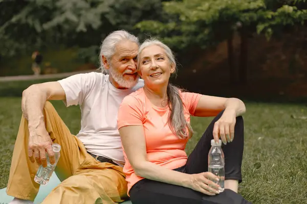 Senior man and woman with yoga mats sitting at park with bottles of water. Positive mature man and his elderly wife after fitness or yoga class in nature. Wellness and healthy lifestyle on retirement.