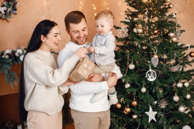 Genç bir aile Noel 'i birlikte kutluyor. Esmer anne, sakallı baba ve sarışın oğul Noel ağacının yanında duruyor ve bir hediye kutusu tutuyorlar. Bej örgülü giysiler giyen bir aile..