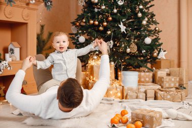 Genç bir aile Noel 'i birlikte kutluyor. Baba oğluyla Noel ağacının yanında eğleniyor. Bej örgülü giysiler giyen bir aile..