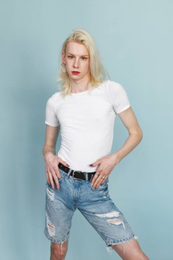 Makyajlı genç sarışın bir adam fotoğraf için poz veriyor. Beyaz tişörtlü ve kırmızı dudaklı bir adam. Mavi arkaplanda izole edilmiş bir adam.