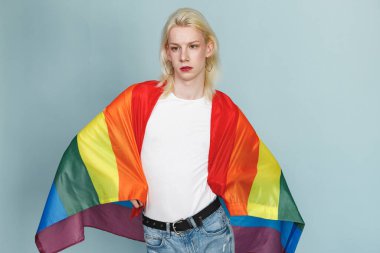 Yüzünde makyaj ve gökkuşağı bayrağı olan genç sarışın bir adam fotoğraf için poz veriyor. Beyaz tişörtlü ve kırmızı dudaklı bir adam. Mavi arkaplanda izole edilmiş bir adam.