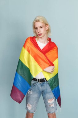 Yüzünde makyaj ve gökkuşağı bayrağı olan genç sarışın bir adam fotoğraf için poz veriyor. Beyaz tişörtlü ve kırmızı dudaklı bir adam. Mavi arkaplanda izole edilmiş bir adam.