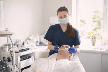 Adam kozmetolog kabinesinde yatıyor ve yüz derisi için bir prosedürü var. Mavi tıbbi kostüm giyen kadın kozmetik uzmanı. Erkeklerin yüzünü pamukla silen kadın..