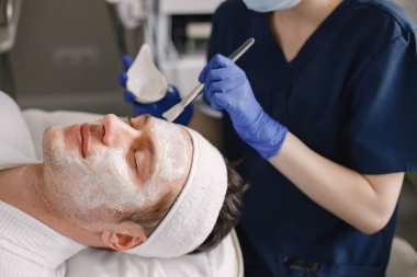 Adam kozmetolog kabinesinde yatıyor ve yüz derisi için bir prosedürü var. Mavi tıbbi kostüm giyen tanınmamış bir kadın kozmetik uzmanı. Erkek yüzüne maske takan bir kadın..