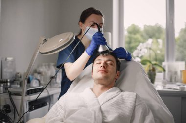 Kozmetolog kabinesinde yatan genç bir adam ve yüz derisi için bir çürüme prosedürü var. Mavi tıbbi kostüm ve yüz maskesi takan kadın kozmetik uzmanı. Beyaz bornoz giyen adam..