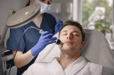 Kozmetolog kabinesinde yatan genç bir adam ve yüz derisi için bir çürüme prosedürü var. Mavi tıbbi kostüm giyen kadın kozmetik uzmanı. Beyaz bornoz giyen adam..