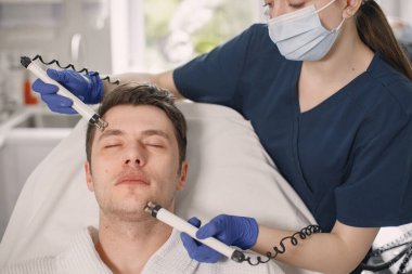 Kozmetolog kabinesinde yatan genç bir adam ve yüz derisi için bir çürüme prosedürü var. Mavi tıbbi kostüm ve yüz maskesi takan kadın kozmetik uzmanı. Beyaz bornoz giyen adam..