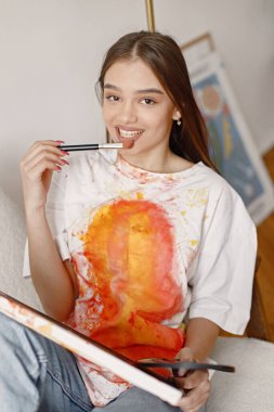 Fırça tutarken ve resim yaparken poz veren genç bir kadın sanatçı. Beyaz tişört ve kot pantolon giyen bir kadın. Beyaz duvarlı bir sandalyede oturan esmer kız..