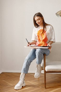 Fırça tutarken ve resim yaparken poz veren genç bir kadın sanatçı. Beyaz tişört ve kot pantolon giyen bir kadın. Beyaz duvarlı bir sandalyede oturan esmer kız..