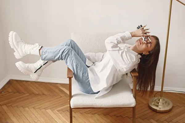 Satisfied female artist posing holding brushes for make up in hands. Woman wearing jeans and white shirt. Brunette girl sitting on a chair in room with white wall.