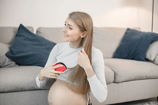 Portrait of pregnant woman brushing her hair. Pregnant woman sitting on a sofa in living room. Blonde woman wearing grey sweater.
