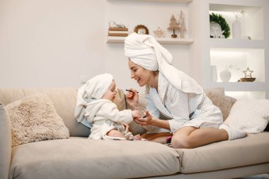Genç anne ve küçük kız havluya sarılı sabahlıklar giyiyorlar. Kanepede oturan kadın ve kız. Elinde dekoratif kozmetik ürünleri tutan kadın..