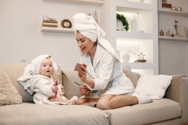Genç anne ve küçük kız havluya sarılı sabahlıklar giyiyorlar. Kanepede oturan kadın ve kız. Elinde dekoratif kozmetik ürünleri tutan kadın..